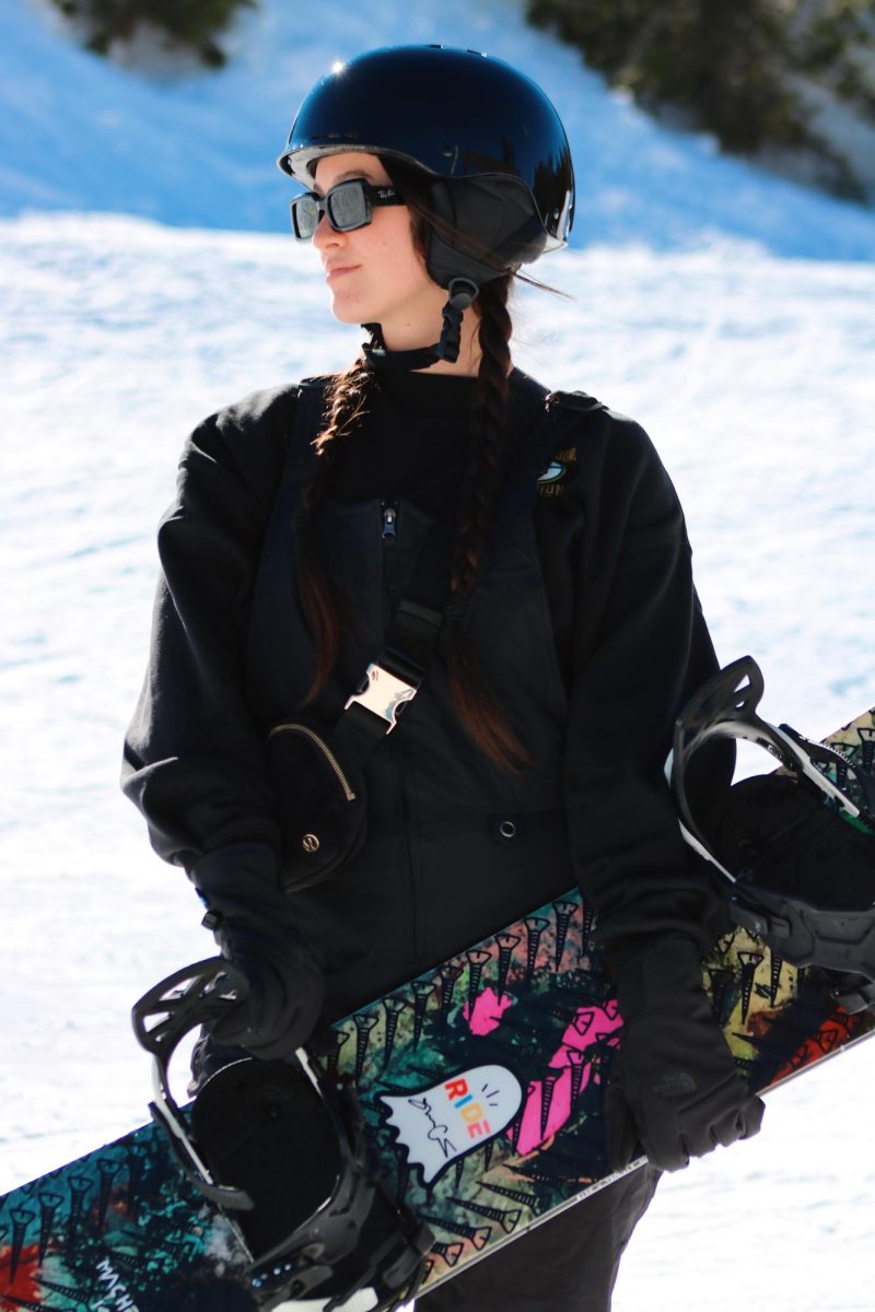 A single shot of a snowboarder holding her board, sunglasses on and ready to start the day of snowboarding.
